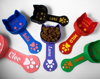 May include: Colorful cat-shaped pet food scoops in various colors, including red, blue, green, and purple. Each scoop has a paw print design and a name, such as "Bella" and "Milo". The scoops are designed to measure pet food.