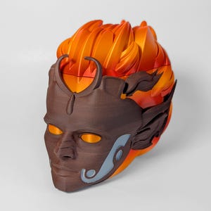 May include: A 3D-printed mask with a stylized face and flame-like hair. The mask is primarily brown with orange and yellow accents, and features a detailed design with a smooth finish. The eyes glow with an orange hue, adding to the mask's dramatic appearance.