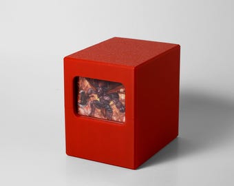 May include: A bright red, cube-shaped card storage box. The box has a textured top and a rectangular window displaying a fantasy illustration. The illustration features a muscular figure with a weapon, set against a fiery background.