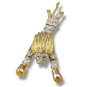 Op de afbeelding: Een goudkleurige metalen broche in de vorm van een gestileerde leeuw, met heldere kristallen accenten en groene edelsteenogen. De leeuw is afgebeeld in een dynamische pose, met uitgestrekte poten. De broche is een vintage stuk.