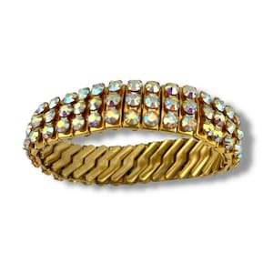Op de afbeelding: Goudkleurige stretcharmband met drie rijen iriserende, ronde strass-steentjes. De armband heeft een zigzagontwerp op de band. De strass-steentjes reflecteren meerdere kleuren, waardoor een sprankelend effect ontstaat. Op een witte achtergrond.