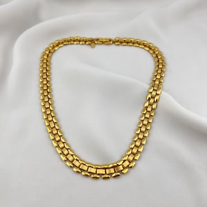 Può includere: Una collana color oro con un design a doppia fila. La collana presenta una serie di maglie rettangolari, creando un aspetto strutturato. La chiusura è visibile in alto e la collana poggia su un tessuto bianco drappeggiato.
