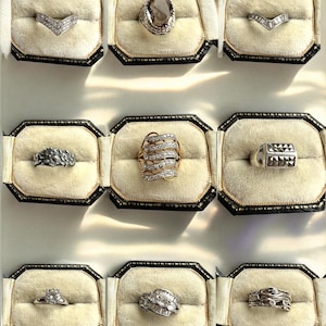 Peut inclure: Neuf bagues présentées dans des boîtes individuelles en velours noir et crème. Les bagues présentent divers modèles, dont une bague en forme de V, une bague avec une grosse pierre ovale et des bagues avec plusieurs petites pierres.
