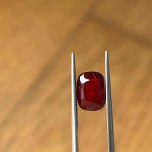 Op de afbeelding: Een kussenvormige, dieprode edelsteen wordt vastgehouden door een zilveren pincet tegen een vage, lichtbruine achtergrond. De facetten van de edelsteen reflecteren licht, wat de helderheid en kleur laat zien. Deze afbeelding benadrukt de schoonheid van een edelsteen.