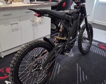 May include: Black electric dirt bike with knobby tires, a black seat, and a gold-colored suspension. The bike is parked on a black and gray mat with a red logo. A second bike is partially visible in the background.