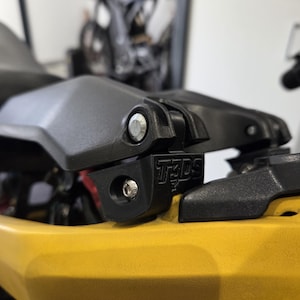 Peut inclure: Gros plan sur un composant noir de moto avec le texte "T3DS" visible. Le composant est fixé à un cadre jaune avec un boulon argenté. Une moto floue est en arrière-plan.