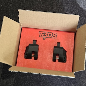 Peut inclure: Une boîte en carton ouverte révèle une boîte rouge avec le texte blanc "T3DS". À l'intérieur, deux composants métalliques noirs sont visibles. Les composants ont une forme rectangulaire avec un dessus saillant et une goupille argentée.