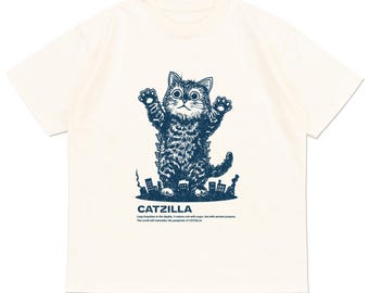 May include: Cream-colored t-shirt featuring a navy blue illustration of a cat in a Godzilla-like pose, with the text "CATZILLA" and additional text below. The cat is depicted with its arms raised, standing in front of a cityscape.