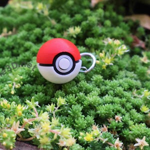 May include: A red, white, and black Poké Ball charm with a silver-colored loop, resting on a bed of green plants with small yellow flowers. The charm is a miniature replica of the iconic ball from the popular animated series.
