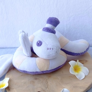 Peut inclure: Une peluche en forme de serpent aux tons blancs, lavande et pêche, coiffée d'un petit chapeau. Le serpent est enroulé sur une surface en bois, ornée de fleurs blanches et jaunes. Le jouet présente des détails brodés et une expression amicale.