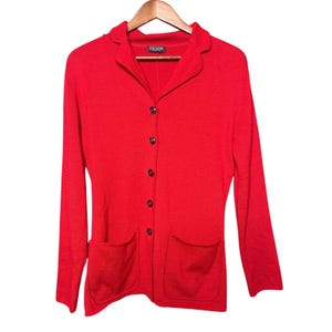 May include: A bright red long-sleeved cardigan featuring a collared neckline, a front button closure with five matching buttons, and two large patch pockets at the bottom. The interior label at the neck displays the brand name ESCADA.