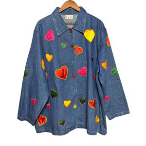 Puede incluir: Chaqueta vaquera azul con aplicaciones de corazones coloridos en rojo, rosa, amarillo y verde. La chaqueta tiene cuello, mangas largas y cierre de botones. La etiqueta muestra "Karen Hart".
