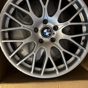 Pode incluir: Uma roda de carro de liga de prata com um design de raios múltiplos e um logotipo da BMW no centro. A roda está em uma caixa de papelão.
