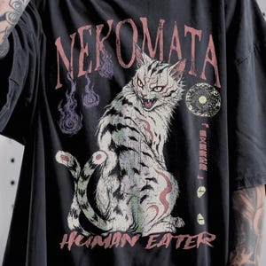 May include: A black short-sleeve t-shirt featuring a graphic print of a two-tailed cat with bared teeth. The text "NEKOMATA" appears in red above the cat, and "HUMAN EATER" is printed in red below. Additional Japanese text and purple flames are visible.
