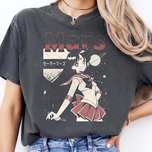 May include: Dark gray t-shirt featuring a retro graphic of a Sailor Mars anime character with the word "Mars" above. The design includes Japanese text and a red and white color scheme. The shirt is paired with blue jeans.