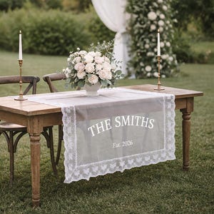 May include: A wooden table set for a wedding or event, with a floral arrangement, candles, and a decorative runner. The runner has the text "THE SMITHS Est. 2026" in white. The table is set outdoors.