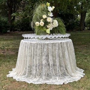 Op de afbeelding: Een ronde tafel bedekt met een crèmekleurig kanten tafelkleed dat tot op de grond reikt met een bloemmotief. Op de tafel staat een rij heldere glazen coupes in een cirkel, rondom een groot bloemstuk van witte rozen.