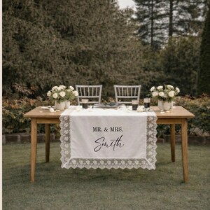 Puede incluir: Un camino de mesa rectangular blanco con un intrincado borde de encaje. El centro del camino presenta el texto impreso en negro "MR. & MRS." sobre el nombre "Smith" en una fuente cursiva, colocado sobre una mesa de madera.