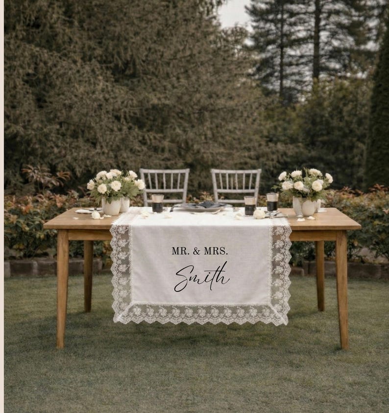 Puede incluir: Un camino de mesa rectangular blanco con un intrincado borde de encaje. El centro del camino presenta el texto impreso en negro "MR. & MRS." sobre el nombre "Smith" en una fuente cursiva, colocado sobre una mesa de madera.