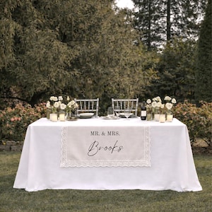 Puede incluir: Un camino de mesa rectangular de tela beige con borde de encaje blanco. El camino presenta el texto impreso MR. & MRS. sobre el nombre Brooks en una fuente cursiva negra, centrado sobre un mantel blanco con cubiertos y vajilla.