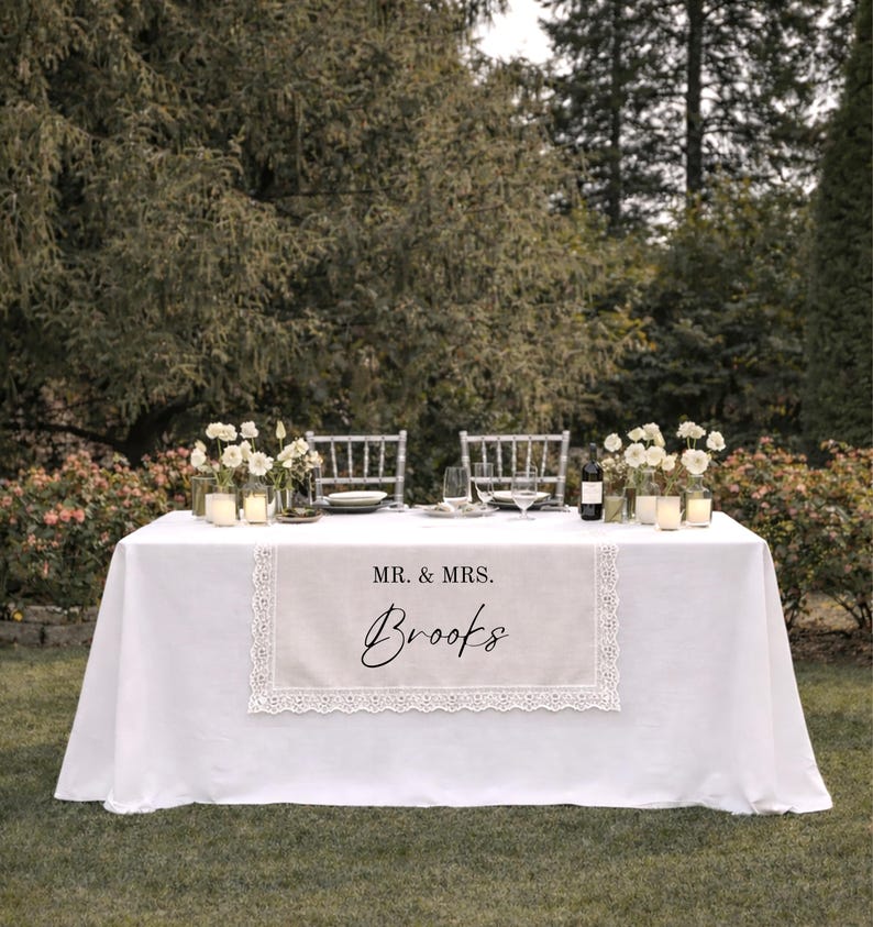 Puede incluir: Un camino de mesa rectangular de tela beige con borde de encaje blanco. El camino presenta el texto impreso MR. & MRS. sobre el nombre Brooks en una fuente cursiva negra, centrado sobre un mantel blanco con cubiertos y vajilla.