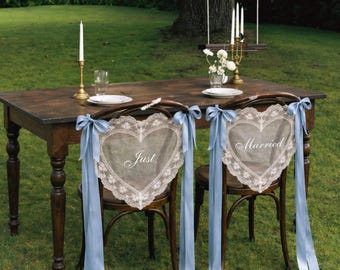 May include: Two wooden chairs featuring heart-shaped white lace covers with the text Just Married in white script. Each chair is adorned with long, light blue satin ribbons tied in bows at the top corners, positioned at a dark wooden table.