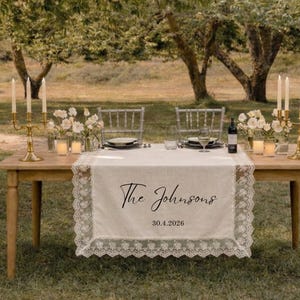 Puede incluir: Una mesa rectangular de madera con un camino de mesa de tela color crema con borde de encaje blanco. El camino tiene impreso el texto The Johnsons y la fecha 30.4.2026 en negro. Dos candelabros dorados con velas blancas decoran la mesa.
