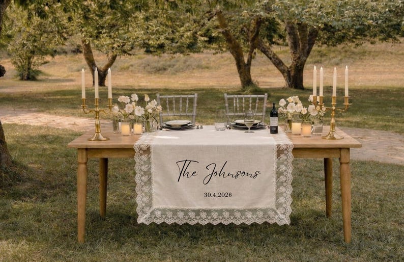 Puede incluir: Una mesa rectangular de madera con un camino de mesa de tela color crema con borde de encaje blanco. El camino tiene impreso el texto The Johnsons y la fecha 30.4.2026 en negro. Dos candelabros dorados con velas blancas decoran la mesa.