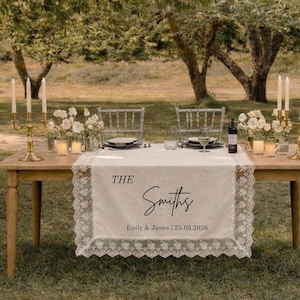 Puede incluir: Un camino de mesa rectangular de color blanco roto con bordes de encaje intrincados, que presenta el texto impreso THE Smiths y Emily & James | 25.03.2026. El camino cubre una mesa de madera con candelabros dorados, flores y cristaler&iacute;a.
