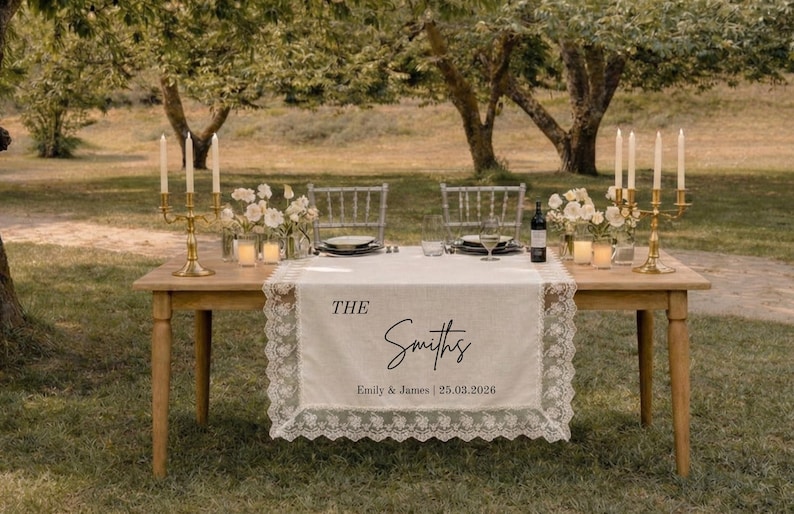 Puede incluir: Un camino de mesa rectangular de color blanco roto con bordes de encaje intrincados, que presenta el texto impreso THE Smiths y Emily & James | 25.03.2026. El camino cubre una mesa de madera con candelabros dorados, flores y cristaler&iacute;a.