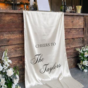 May include: A long, cream-colored satin table runner draped over a wooden bar. The fabric features black printed text that reads "CHEERS TO" in a simple sans-serif font, followed by "The Taylors" in a decorative, elegant cursive script.