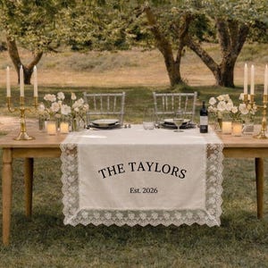 Könnte beinhalten: Ein Holztisch für eine Hochzeitsfeier im Freien. Der Tisch ist mit einem weißen Spitzentischläufer mit dem Text "THE TAYLORS Est. 2026" dekoriert. Goldene Kerzenhalter, Blumen und transparente Stühle vervollständigen die elegante Einrichtung.