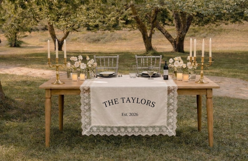 Puede incluir: Un camino de mesa rectangular de color blanco roto con un borde de encaje blanco intrincado. El centro presenta un texto impreso en negro que dice THE TAYLORS y Est. 2026. El camino se muestra sobre una mesa de madera con candelabros dorados.