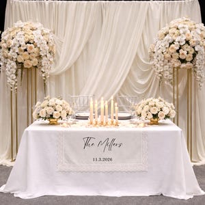 Puede incluir: Un camino de mesa blanco con borde de encaje que presenta el texto impreso en negro The Millers y la fecha 11.3.2026. El camino est&aacute; colocado sobre un mantel blanco con un centro de mesa de velas, arreglos florales y un fondo de tela drapeada.