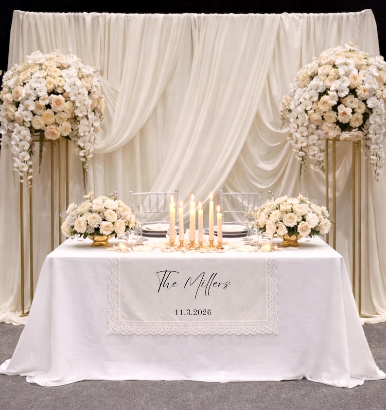 Puede incluir: Un camino de mesa blanco con borde de encaje que presenta el texto impreso en negro The Millers y la fecha 11.3.2026. El camino est&aacute; colocado sobre un mantel blanco con un centro de mesa de velas, arreglos florales y un fondo de tela drapeada.