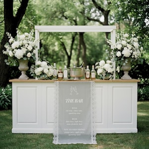 Può includere: Un bancone in legno bianco con piano in legno chiaro, dotato di due grandi urne in pietra riempite di fiori bianchi e fogliame. Un runner traslucido con il testo THE BAR, WINE, BEER e NON ALCOHOL DRINKS pende dal centro, con bottiglie e un secchiello.