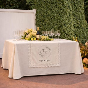 Puede incluir: Un mantel rectangular de color crema con un camino de mesa central con borde de encaje. El camino muestra un dise&ntilde;o floral circular con las iniciales TD y el texto Tayla & Dylan. Una botella de vino, copas y limas verdes sobre la mesa.