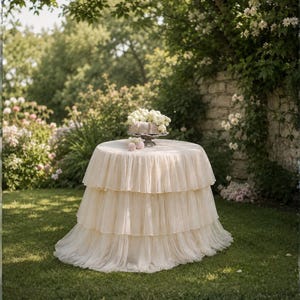 May include: A round table covered with a cream-colored, tiered, ruffled fabric. The table is set on a green lawn, with a cake and flowers on top. The background features a garden setting with trees and a stone wall.