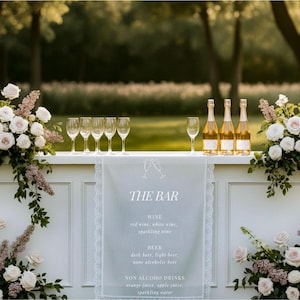 May include: A white bar decorated with floral arrangements and champagne bottles. The bar has a sign that reads "THE BAR" with a list of drinks. Wine glasses are arranged on the bar.