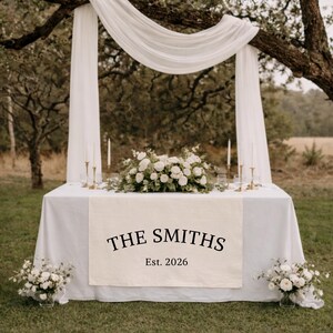 Puede incluir: Un camino de mesa rectangular blanco con el texto impreso en negro THE SMITHS y Est. 2026, sobre un mantel blanco. La mesa est&aacute; decorada con un arreglo floral blanco central, cuatro velas finas y dos ramos de flores a los lados.