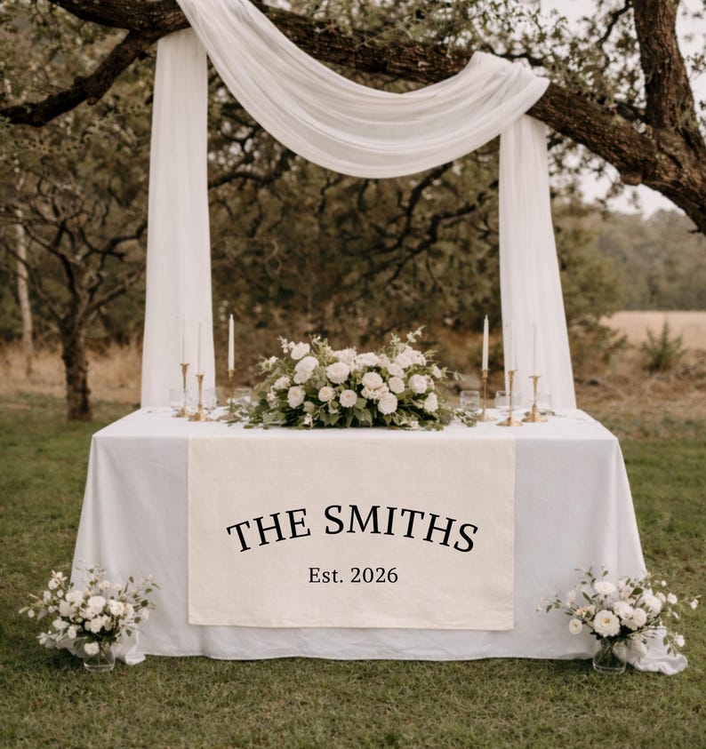 Puede incluir: Un camino de mesa rectangular blanco con el texto impreso en negro THE SMITHS y Est. 2026, sobre un mantel blanco. La mesa est&aacute; decorada con un arreglo floral blanco central, cuatro velas finas y dos ramos de flores a los lados.