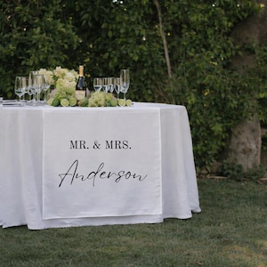 Puede incluir: Un camino de mesa de tela blanca sobre una mesa redonda, con el texto impreso en negro MR. & MRS. Anderson. La mesa est&aacute; dispuesta con varias copas de cristal transparente, una botella de champ&aacute;n y un centro de limas verdes y flores blancas.
