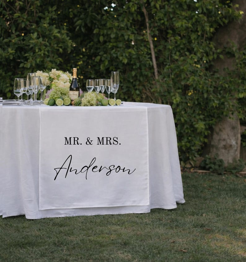 Puede incluir: Un camino de mesa de tela blanca sobre una mesa redonda, con el texto impreso en negro MR. & MRS. Anderson. La mesa est&aacute; dispuesta con varias copas de cristal transparente, una botella de champ&aacute;n y un centro de limas verdes y flores blancas.
