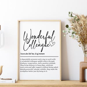 Könnte beinhalten: Gerahmter Druck mit den Worten "Wonderful Colleague" in eleganter Schrift. Die Definition von "colleague" ist ebenfalls enthalten. Eine dekorative Vase mit getrockneten Pflanzen und eine kleine Vogelfigur sind ebenfalls sichtbar.