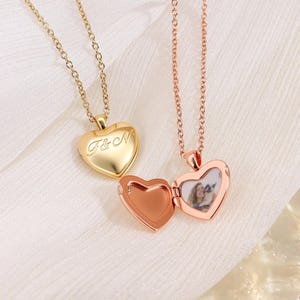 Könnte beinhalten: Zwei herzförmige Medaillon-Halsketten. Eine ist goldfarben mit den Initialen "F&N" auf der Vorderseite eingraviert. Die andere ist roségoldfarben und offen, wodurch sich ein kleines Foto im Inneren zeigt. Beide Halsketten haben zarte Ketten.
