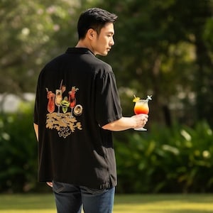 May include: Black short-sleeved shirt with a cocktail design on the back. The shirt features a variety of colorful cocktail illustrations and the word "Soul" in gold. The person is holding a cocktail with a lemon and a straw.