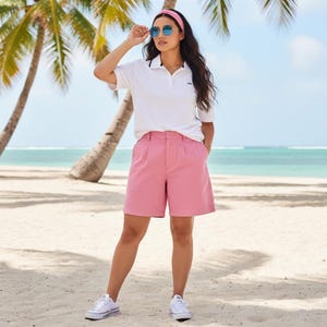 Op de afbeelding: Een vrouw draagt een witte poloshirt met zwarte bies, een roze short, witte sneakers en een roze haarband. Ze draagt een zonnebril en staat op een zandstrand met palmbomen en de oceaan op de achtergrond.