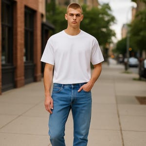May include: A person wearing a white crew neck t-shirt and blue jeans. The person is standing on a sidewalk with a brick building and trees in the background.