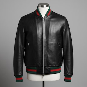 Puede incluir: Chaqueta bomber de cuero negro con cremallera completa. La chaqueta presenta ribetes a rayas verdes y rojas en el cuello, los puños y el dobladillo. La chaqueta se exhibe en un maniquí sobre un fondo gris.