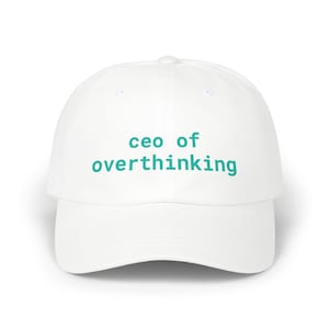 May include: A white baseball cap featuring the text "ceo of overthinking" printed in a teal sans-serif font across the front panels. The cap has a structured crown, a curved brim, and a button at the top center of the dome.